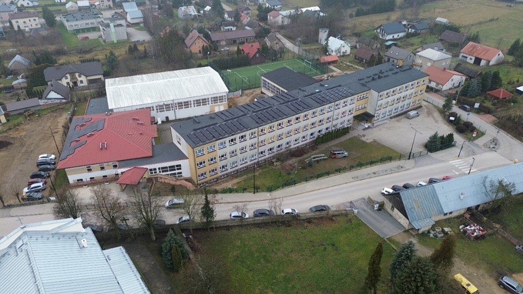 Widok z lotu ptaka na budynek szkoły z panelami fotowoltaicznymi na dachu, salą gimnastyczną i boiskiem