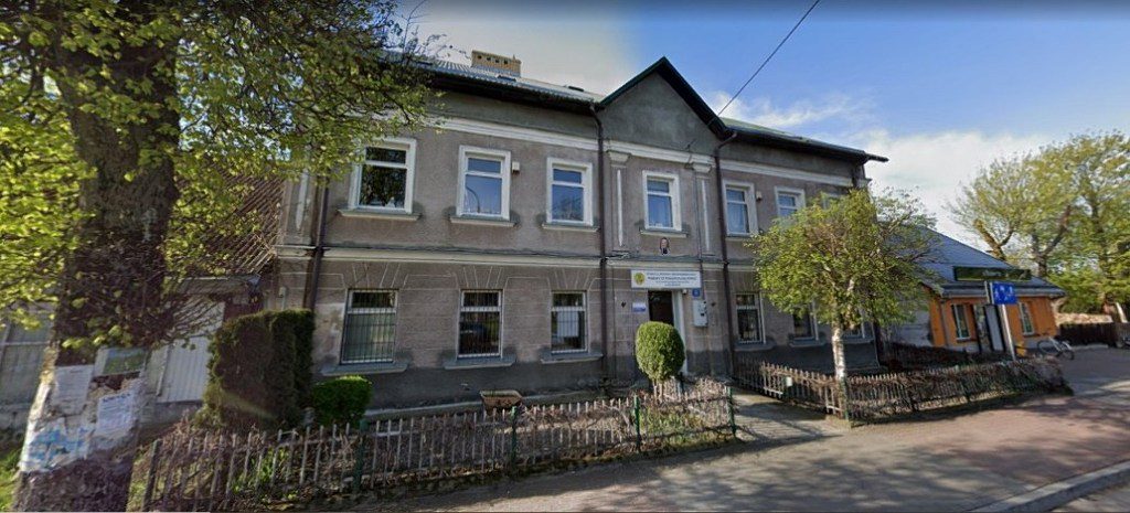 Piętrowy szary budynek z tablicą urzędową nad wejściem, otoczony niskim płotem i drzewami