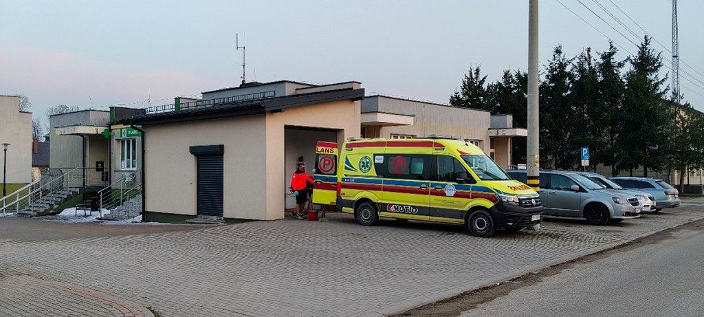 Żółty ambulans zaparkowany przed budynkiem, ratownik medyczny przy otwartych tylnych drzwiach pojazdu