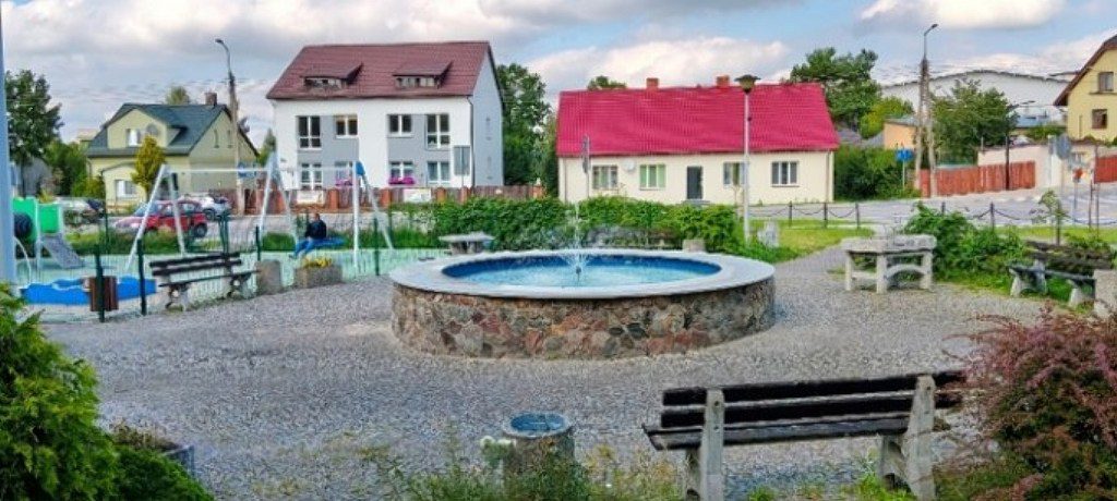 Skwer z okrągłą kamienną fontanną, ławkami i placem zabaw na tle domów jednorodzinnych