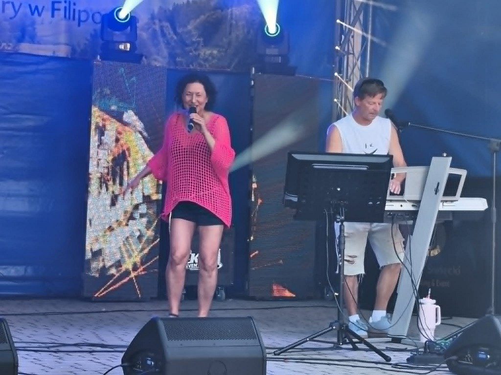 Kobieta śpiewająca do mikrofonu i mężczyzna grający na keyboardzie na scenie plenerowej