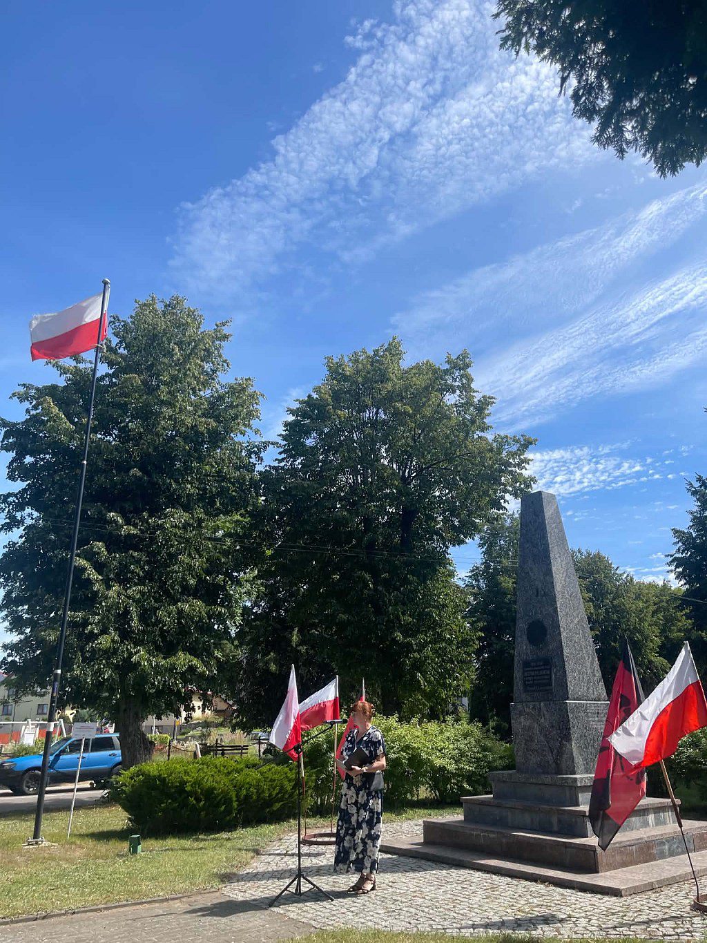 Kobieta stoi przed kamiennym obeliskiem w otoczeniu polskich flag w słoneczny dzień