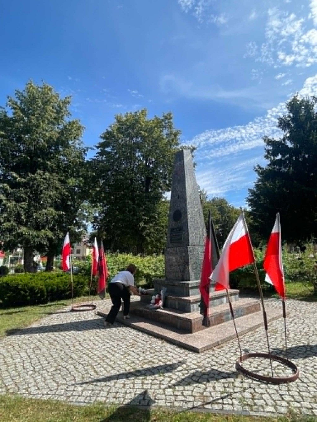 Kobieta składająca kwiaty pod kamiennym obeliskiem w otoczeniu biało-czerwonych flag