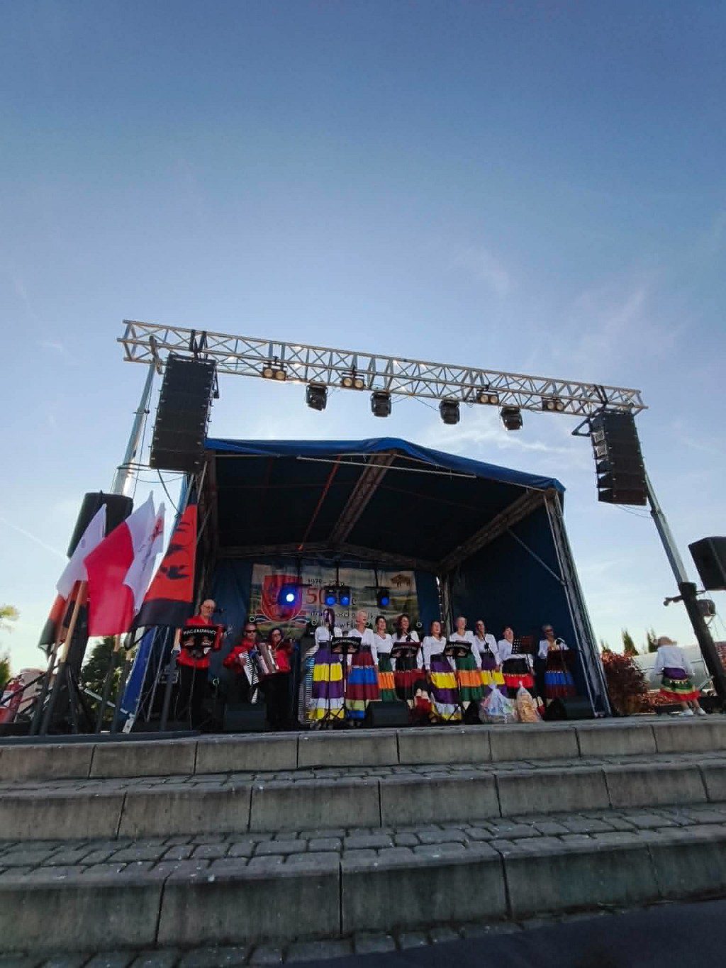 Występ zespołu ludowego w kolorowych strojach na zadaszonej scenie plenerowej podczas lokalnej uroczystości