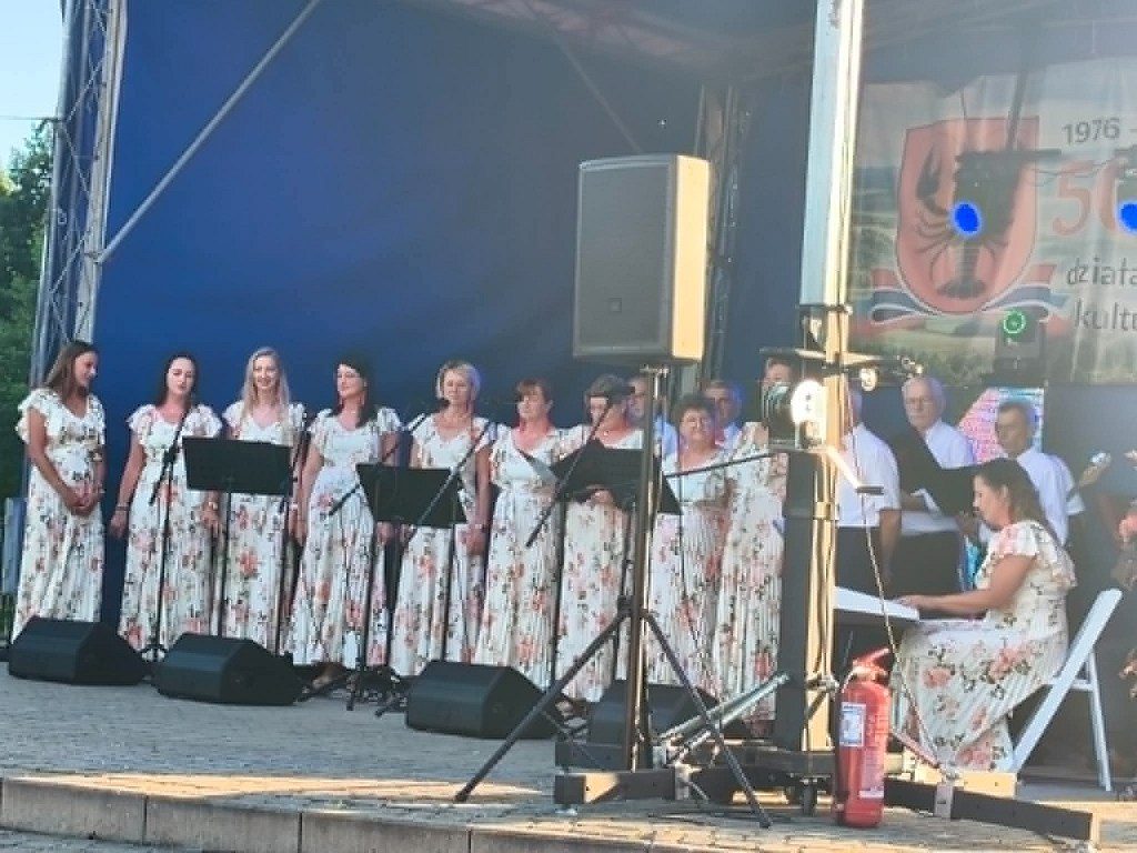 Chór kobiet w kwiecistych sukienkach występujący na scenie plenerowej przy akompaniamencie instrumentu klawiszowego