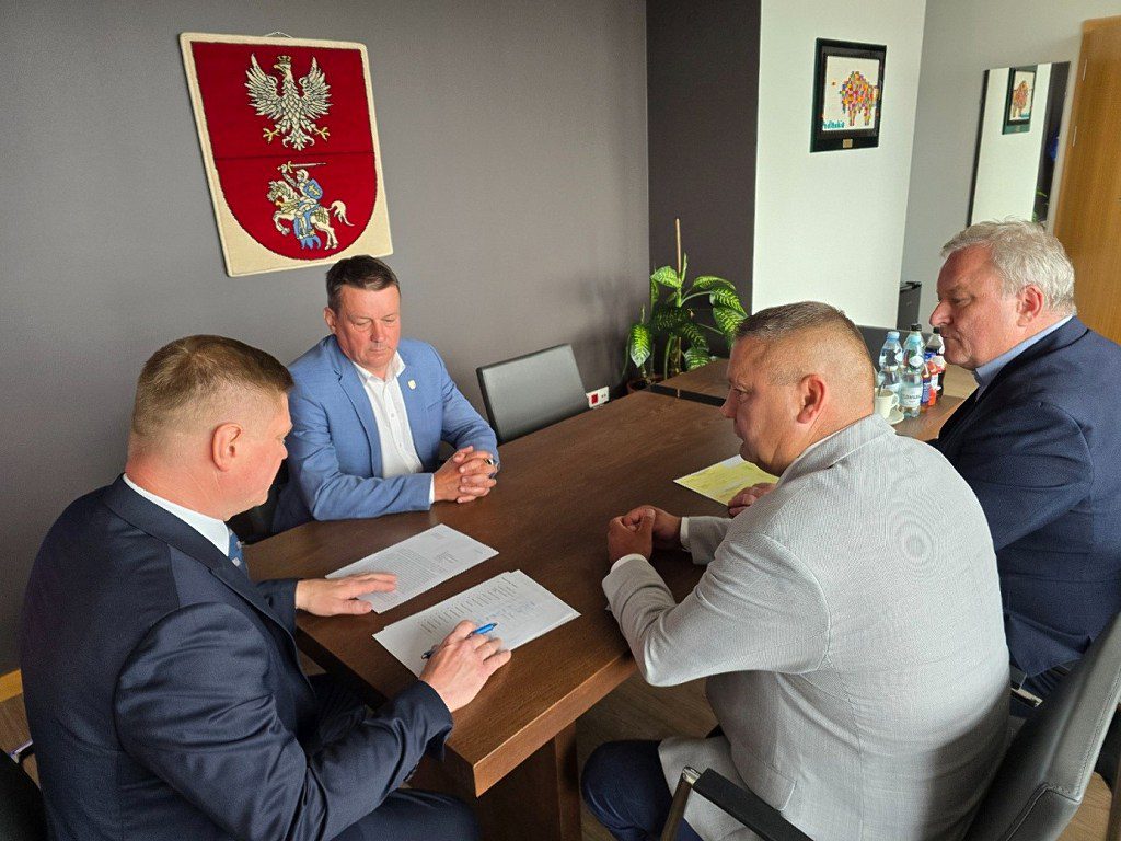 Czterech mężczyzn w garniturach przy stole konferencyjnym z dokumentami, w tle herb województwa podlaskiego