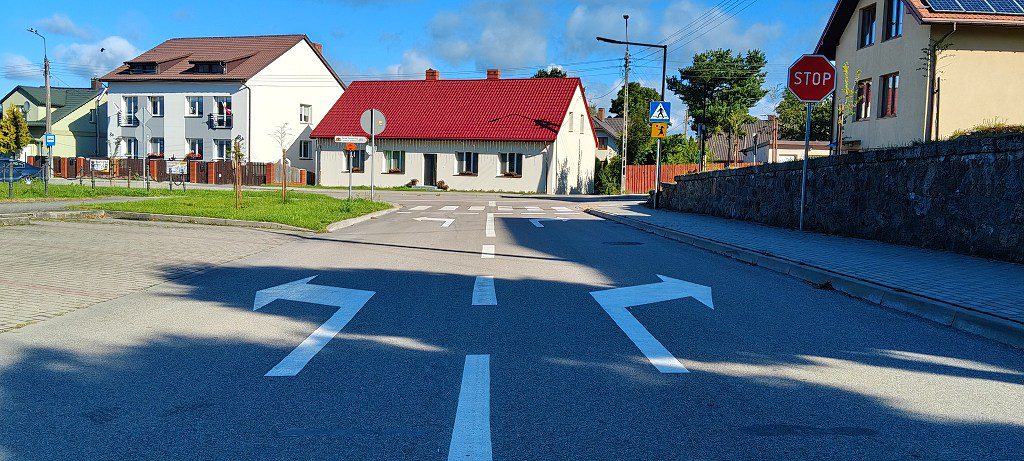 Skrzyżowanie dróg ze strzałkami kierunkowymi na jezdni, znakiem STOP i domami jednorodzinnymi w tle