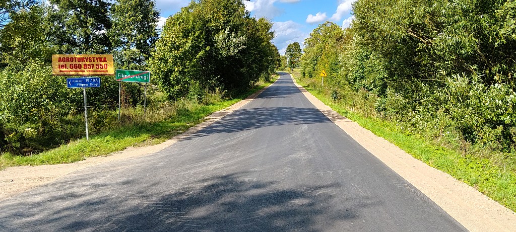Asfaltowa droga otoczona zielenią z tablicą końca miejscowości Filipów Drugi i drogowskazami po lewej stronie