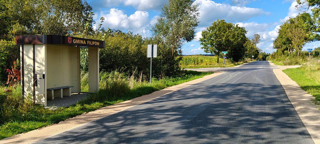 Przystanek autobusowy z napisem Gmina Filipów przy asfaltowej drodze w wiejskim krajobrazie