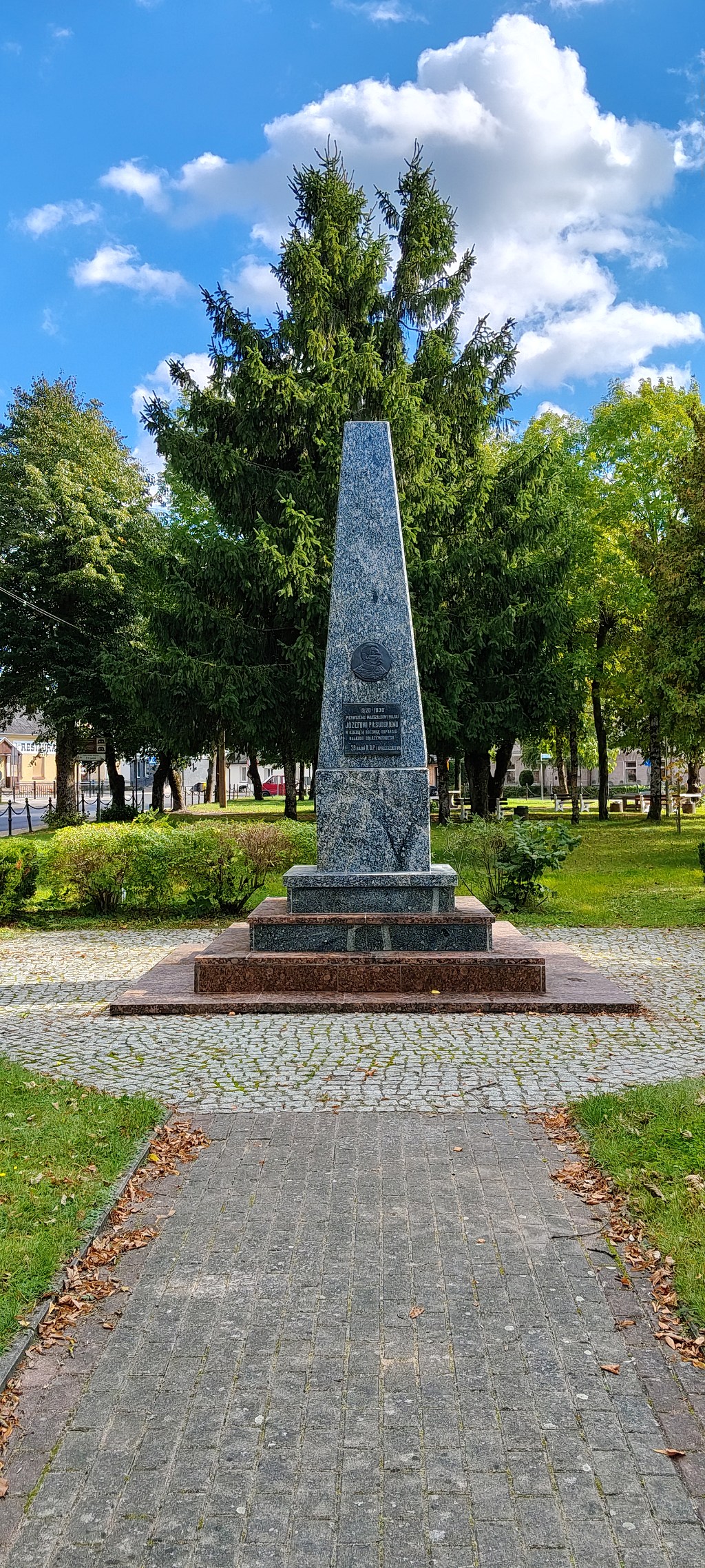 Kamienny obelisk z tablicą pamiątkową dedykowaną Józefowi Piłsudskiemu, usytuowany w parku wśród drzew