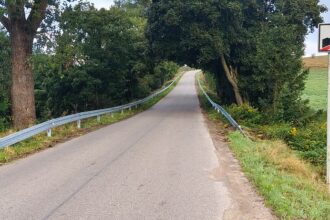 Asfaltowa droga otoczona drzewami, ze znakiem końca obszaru zabudowanego po prawej stronie