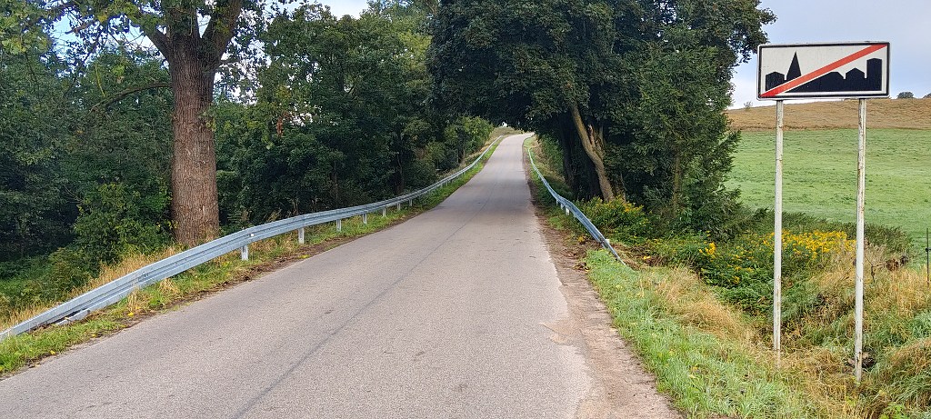 Asfaltowa droga otoczona drzewami, ze znakiem końca obszaru zabudowanego po prawej stronie