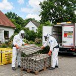 Pracownicy w kombinezonach ochronnych i maskach przenoszą płyty azbestowe przed domem jednorodzinnym