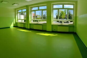 Jasna, pusta sala z zielonymi ścianami, podłogą i dużymi oknami, przez które wpada słońce