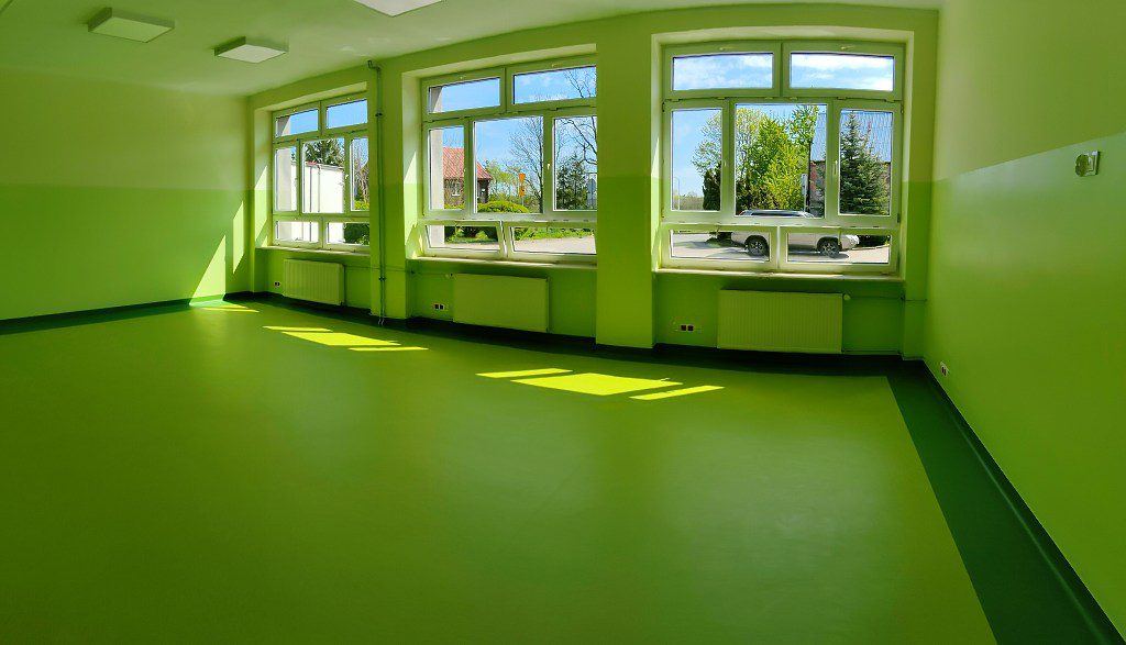 Jasna, pusta sala z zielonymi ścianami, podłogą i dużymi oknami, przez które wpada słońce