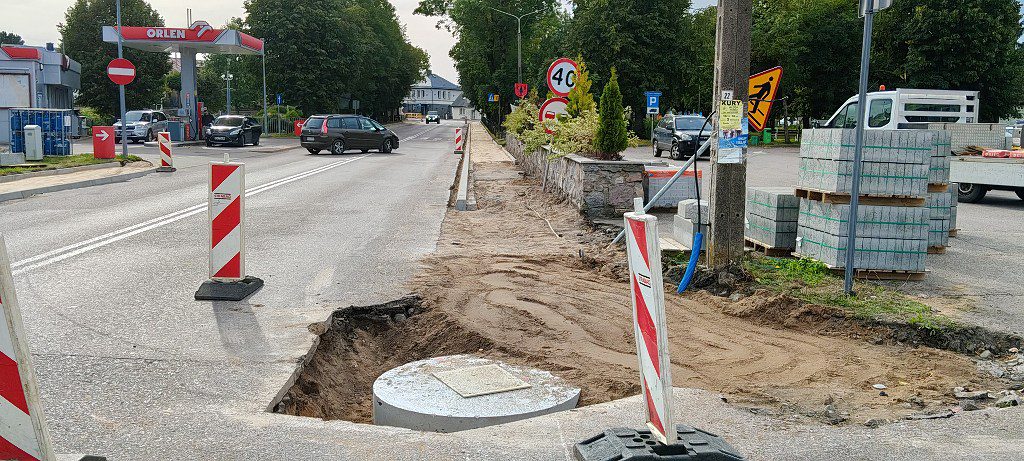 Prace drogowe i remont chodnika przy ulicy, widoczne słupki ostrzegawcze, wykop oraz stacja benzynowa w tle