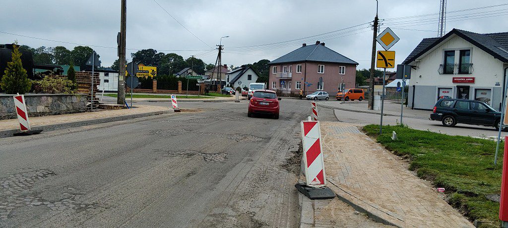 Remont drogi w terenie zabudowanym z czerwono-białymi słupkami ostrzegawczymi i uszkodzoną nawierzchnią