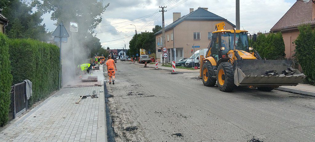 Prace drogowe na ulicy: żółta koparko-ładowarka na jezdni i robotnicy układający kostkę brukową na chodniku