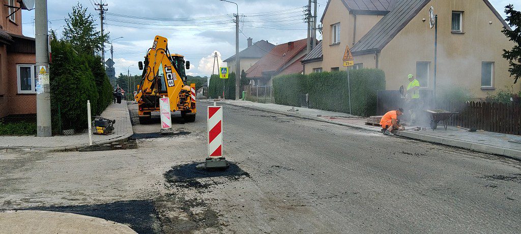 Prace remontowe na drodze osiedlowej z udziałem koparki JCB i robotników układających kostkę brukową