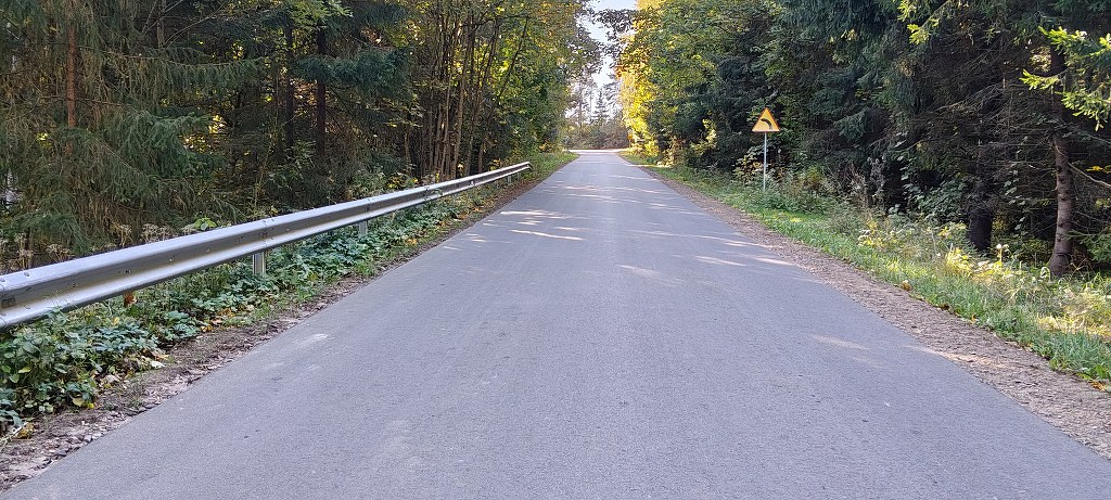 Asfaltowa droga biegnąca przez las z barierką po lewej stronie i znakiem ostrzegawczym przed zakrętem w prawo