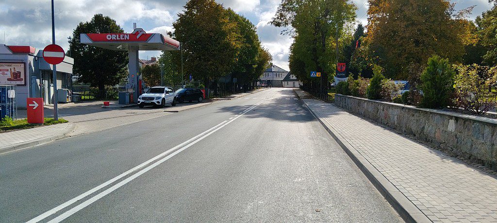 Ulica ze stacją paliw Orlen po lewej stronie, zaparkowanymi samochodami i drzewami wzdłuż drogi