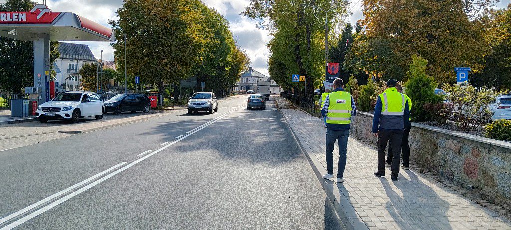 Mężczyźni w żółtych kamizelkach odblaskowych stojący na chodniku przy ulicy obok stacji paliw