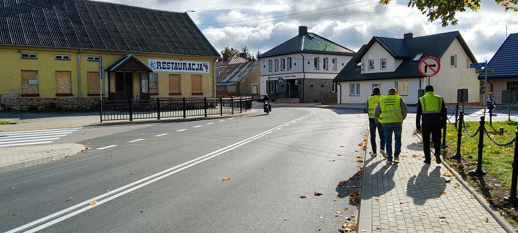 Trzech mężczyzn w żółtych kamizelkach odblaskowych idzie chodnikiem wzdłuż drogi w małej miejscowości