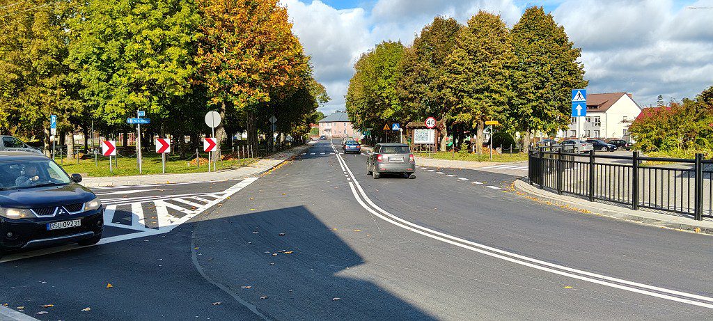 Asfaltowa ulica ze znakami drogowymi, przejściami dla pieszych i jadącymi samochodami, otoczona drzewami