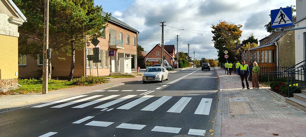 Ulica z nowo namalowanym przejściem dla pieszych, jadącym samochodem i osobami w kamizelkach odblaskowych na chodniku