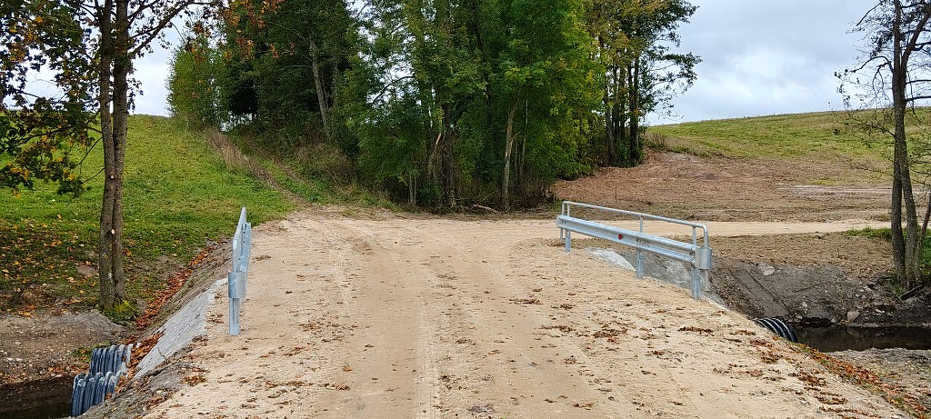 Piaszczysta droga przechodząca przez mały mostek z metalowymi barierkami pośród drzew i pól