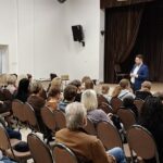 Prelegent prowadzi wykład dla publiczności w sali z wyświetlaną prezentacją na ekranie