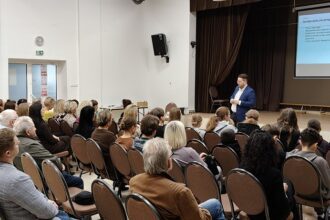 Prelegent prowadzi wykład dla publiczności w sali z wyświetlaną prezentacją na ekranie