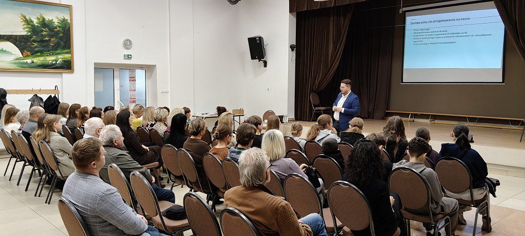 Prelegent prowadzi wykład dla publiczności w sali z wyświetlaną prezentacją na ekranie