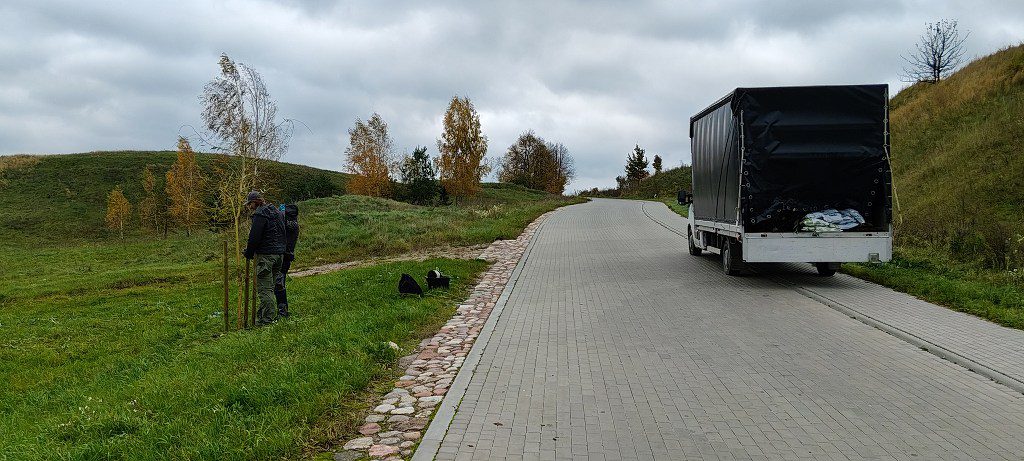 Dwie osoby sadzące młode drzewko na poboczu kostkowanej drogi, obok zaparkowanej ciężarówki