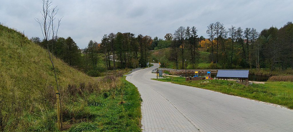 Wybrukowana alejka w parku rekreacyjnym z placem zabaw, drewnianą wiatą i zbiornikiem wodnym wśród zieleni